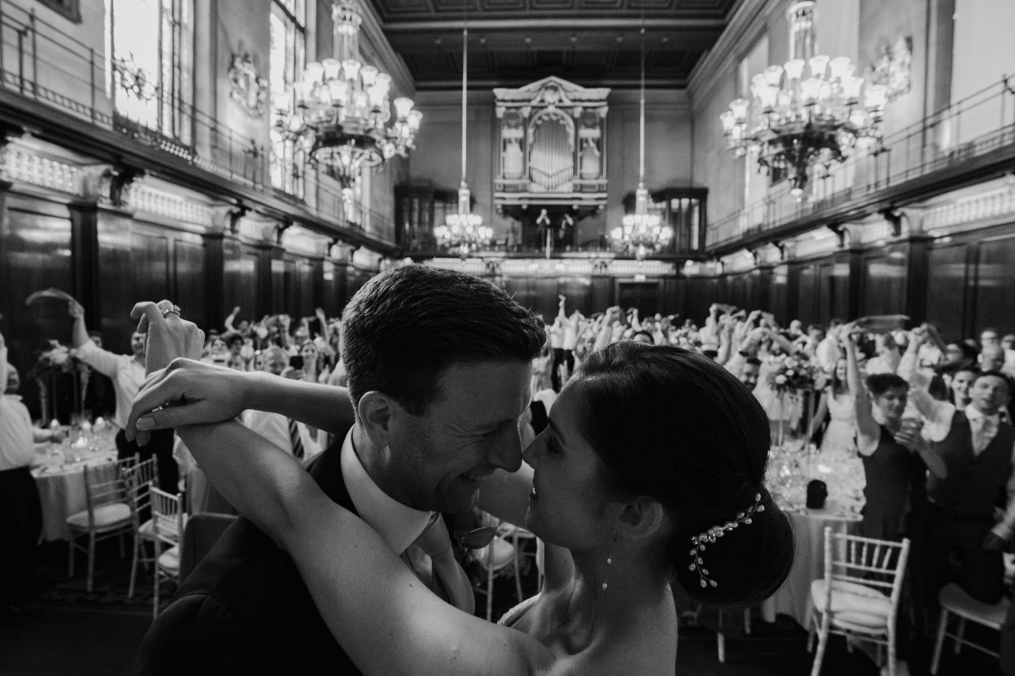 black and white image of bride and groom embracing in front of a crowded room, captured in Merchant Taylors' Hall wedding photography, by Ben Lee