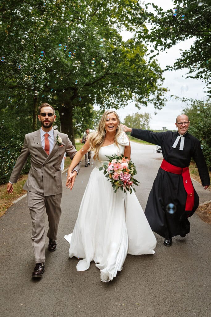 vicar with bubble machine beside bride and groom by Eastfield Barn wedding photographer, Ben Lee.